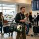 Homem caminhando em terminal de aeroporto com bolsa de viagem verde escura pendurada no corpo.