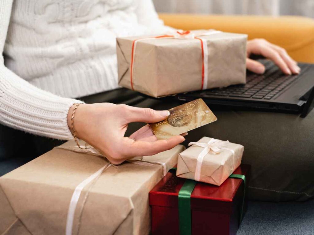 Mulher segurando um cartão de crédito enquanto faz compras online pelo notebook, rodeada por caixas de presentes embrulhadas. A imagem representa uma dona de empresa investindo na compra de itens de qualidade para presentear clientes.