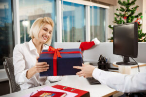 Dando presente de natal para um funcionário em ambiente de trabalho. A funcionária, loira e de camiseta branca, sorri, ao receber uma caixa envelopada em papel de presente azul marinho com fita vermelha