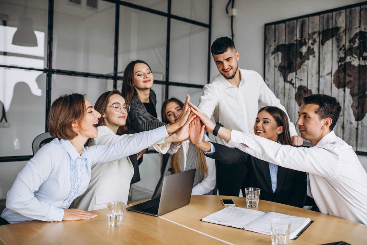 Grupo de profissionais em reunião de trabalho em um escritório, reunidos ao redor de uma mesa e celebrando juntos com um gesto de high five coletivo, transmitindo união e espírito de equipe.