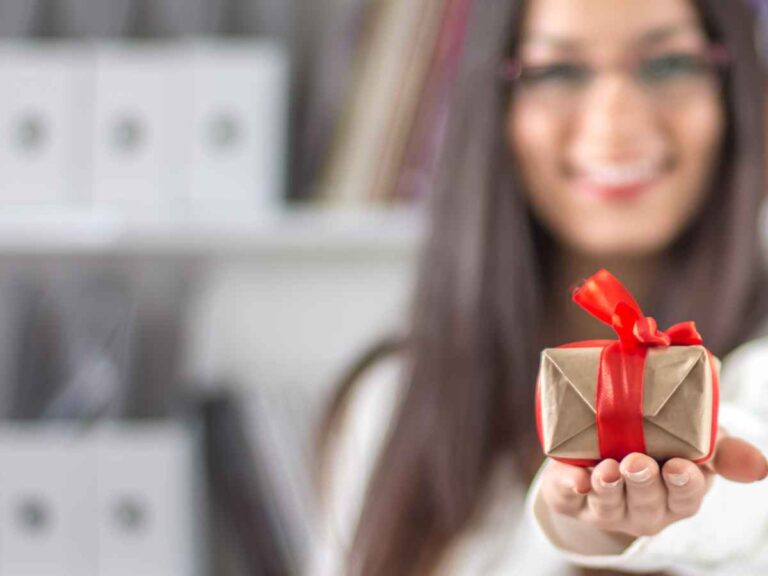 Colaboradora sorridente segurando um presente com laço vermelho, simbolizando a entrega de brindes corporativos personalizados no ambiente de escritório para reconhecimento e engajamento.