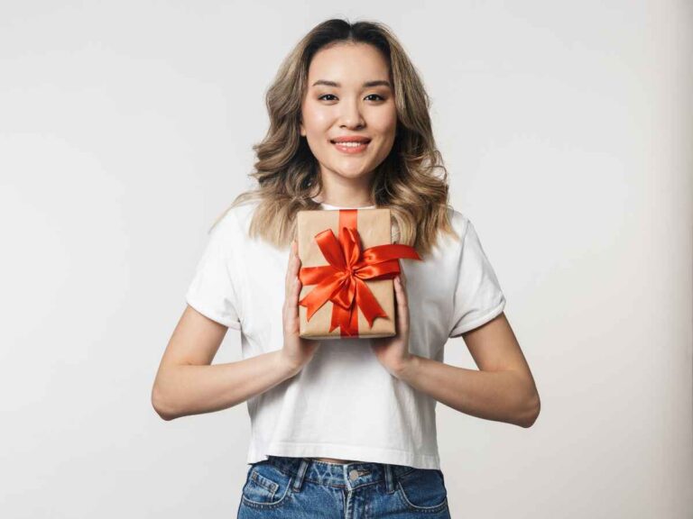 Colaboradora sorridente segurando um brinde. Ela veste blusa branca e calça jeans, segurando o presente embrulhado com um laço vermelho à frente do peito. Fundo branco sólido.