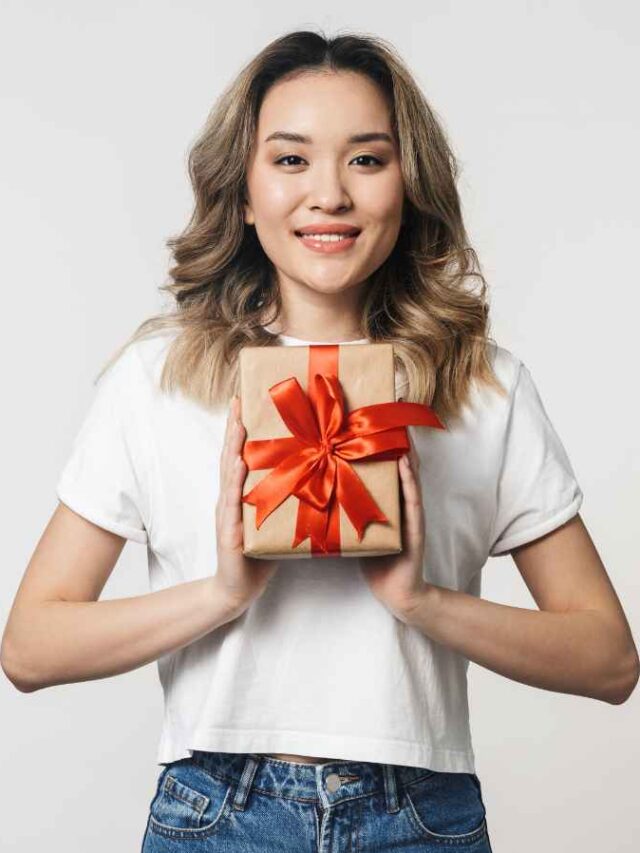 Colaboradora sorridente segurando um brinde. Ela veste blusa branca e calça jeans, segurando o presente embrulhado com um laço vermelho à frente do peito. Fundo branco sólido.