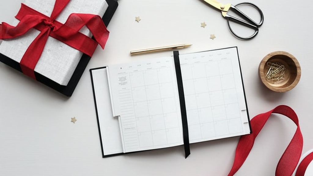 Opção de brindes para funcionários de fim de ano com uma agenda aberta sobre uma mesa, caneta dourada e uma embalagem de presente amarrada com um laço vermelho.