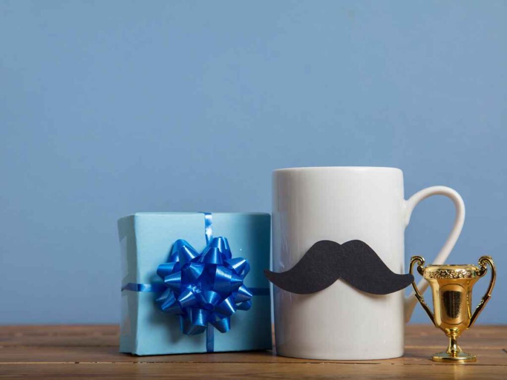 Caneca branca com bigode preto colado, ao lado de uma caixinha azul com laço e um troféu dourado, representando homenagem divertida e carinhosa para o Dia dos Pais.