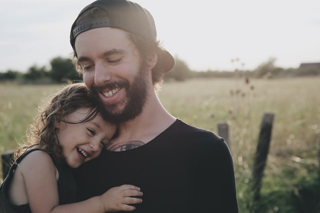 Pai de barba e boné com camiseta preta segurando uma criança, menina, sua filha em seu colo. Os dois sorriem. Atrás, paisagem com grama e árvores