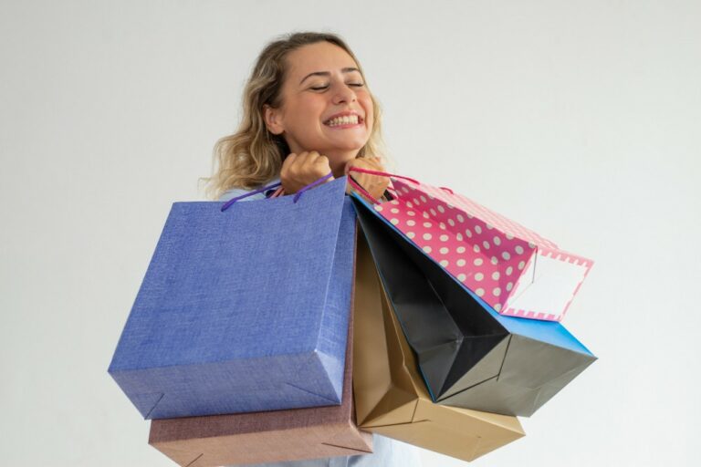 Mulher loira segurando sacolas de brindes para presentear seus clientes em comemoração ao Dia do Colaborador. Fundo branco sólido.