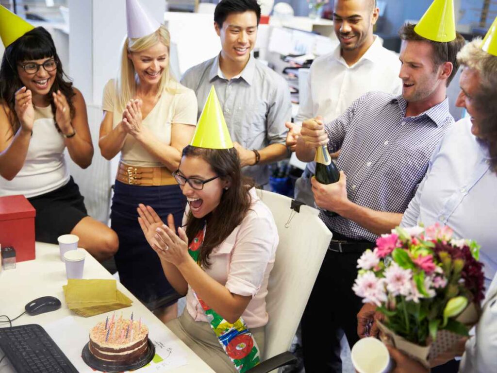 Grupo de colegas de trabalho comemora o aniversário de uma colaboradora no escritório, com bolo, chapéus de festa, flores e champanhe