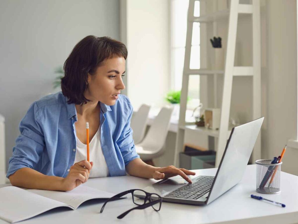 Mulher jovem sentada à mesa de trabalho, usando notebook para estudar, com caderno aberto e lápis na mão, em ambiente iluminado e organizado.