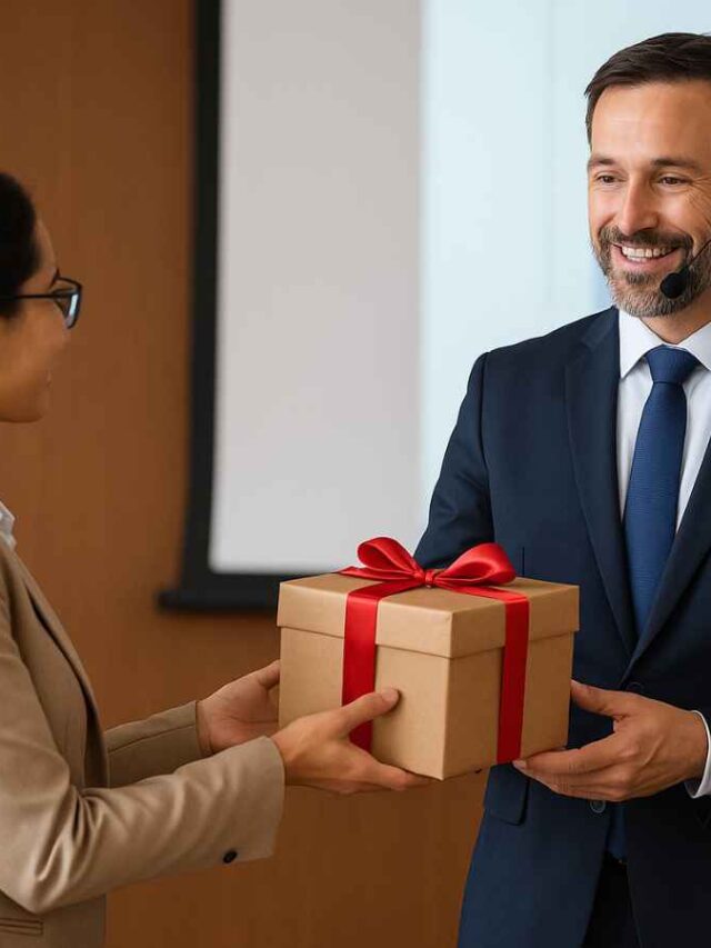 Mulher entregando um presente embrulhado com laço vermelho para um palestrante em um evento corporativo. O palestrante, usando terno e microfone de cabeça, sorri ao receber o brinde como forma de agradecimento pela sua participação e contribuição no evento.