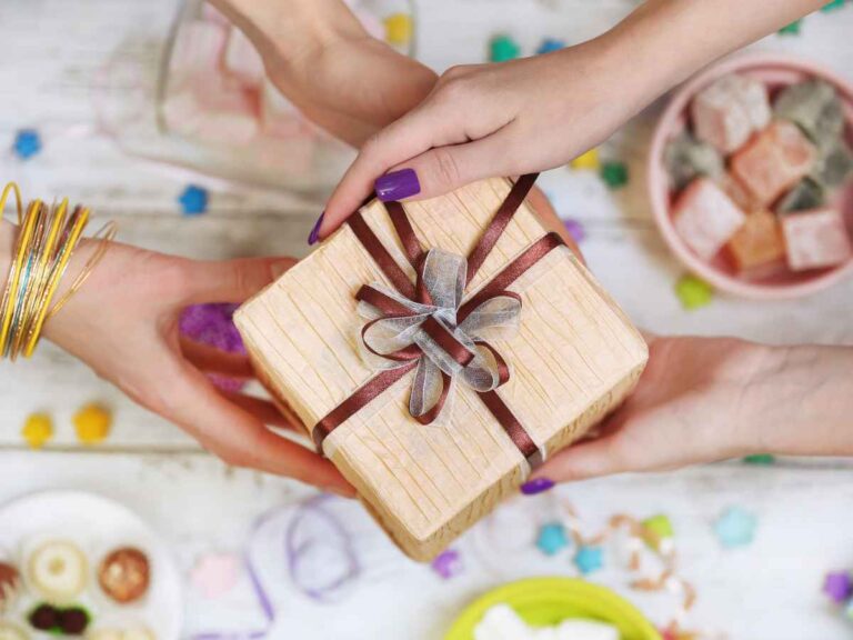 Mãos entregando um presente decorado com laço rústico em meio a uma mesa com doces típicos coloridos, representando um brinde temático de Festa Junina. A imagem remete à entrega de lembranças em um evento corporativo com temática junina, reforçando o clima festivo e o cuidado com a experiência dos colaboradores.