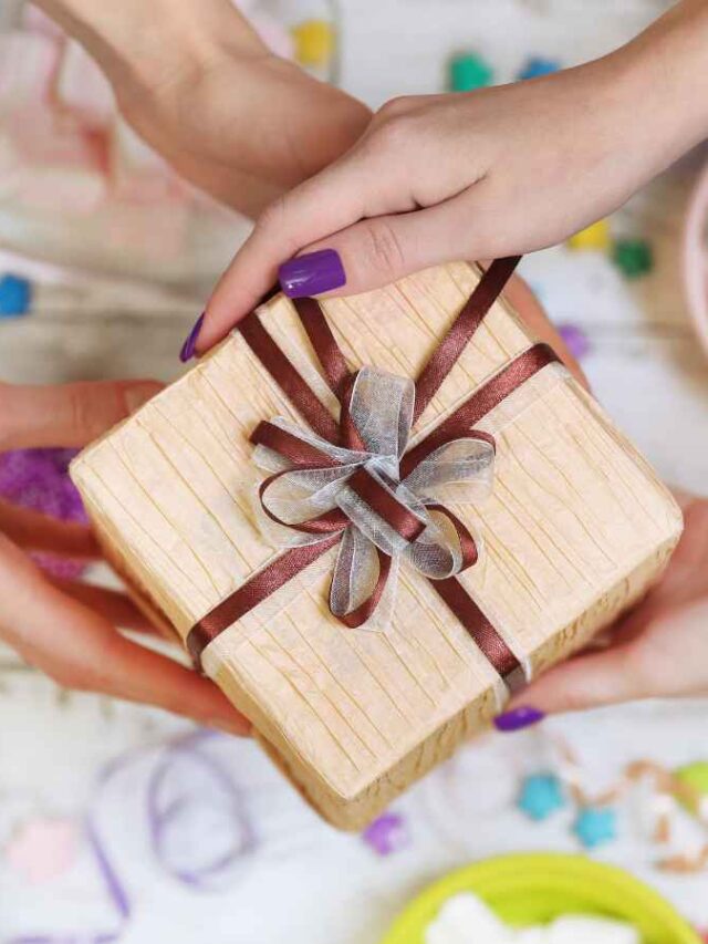 Mãos entregando um presente decorado com laço rústico em meio a uma mesa com doces típicos coloridos, representando um brinde temático de Festa Junina. A imagem remete à entrega de lembranças em um evento corporativo com temática junina, reforçando o clima festivo e o cuidado com a experiência dos colaboradores.