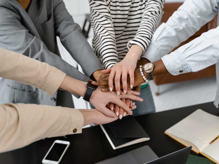 equipe de 4 pessoas juntando as mãos em união durante o trabalho, em cima de uma mesa com cadernos.