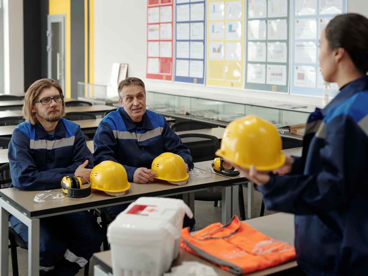 Dois homens vestidos com EPIs, assistindo uma mulher fazer uma apresentação dos equipamentos de segurança e higiene na SIPAT.