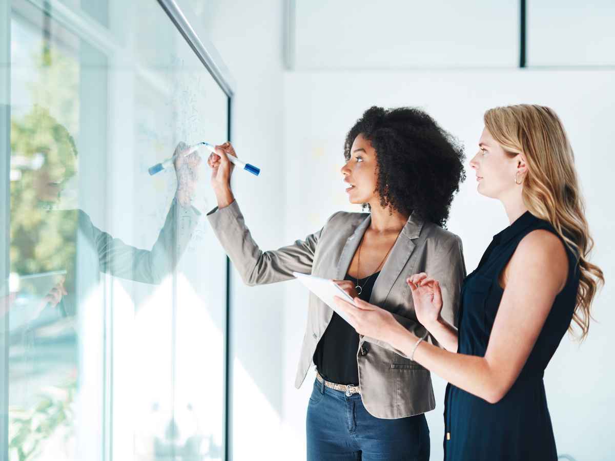 2 mulheres, colaboradoras trabalhando enquanto discutem e planejam escrevendo em uma lousa.