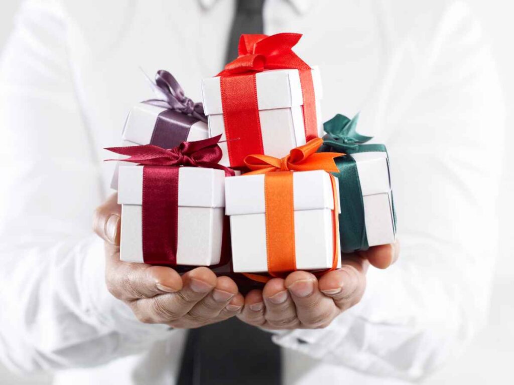 Homem vestindo camisa social branca segurando vários pequenos presentes embrulhados com laços coloridos. A imagem representa brindes personalizados preparados para distribuição em eventos corporativos, destacando a importância de oferecer lembranças criativas e estratégicas para fortalecer a marca e encantar participantes.