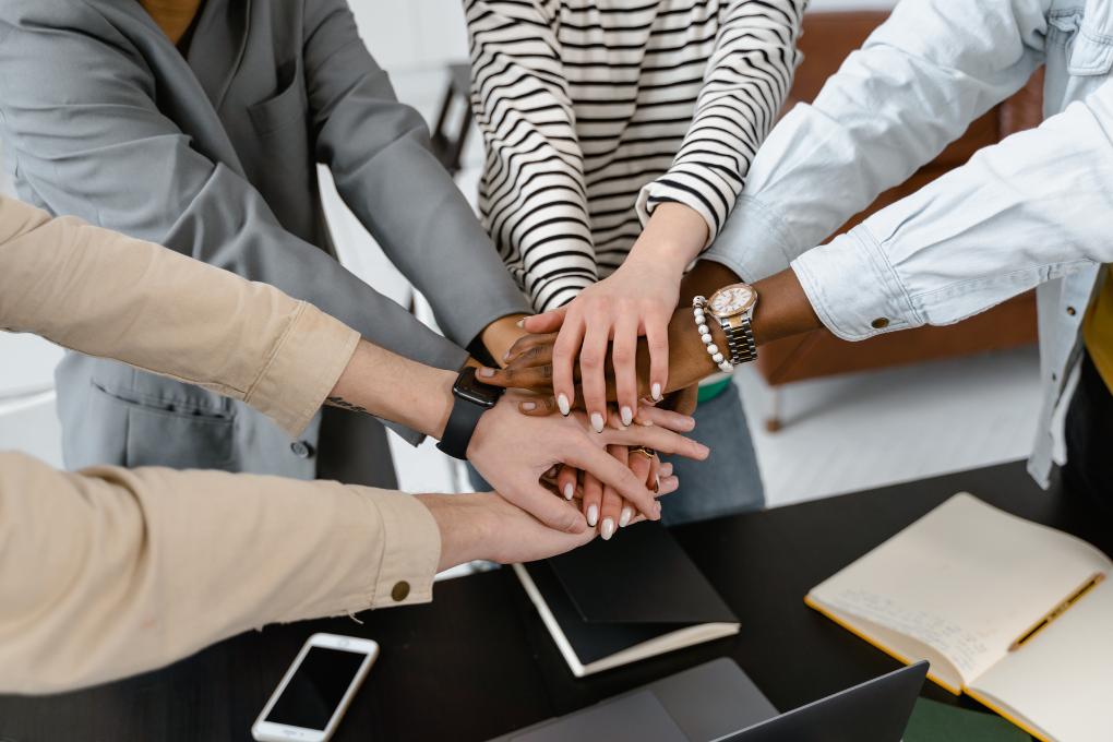 equipe de 4 pessoas juntando as mãos em união durante o trabalho, em cima de uma mesa com cadernos.