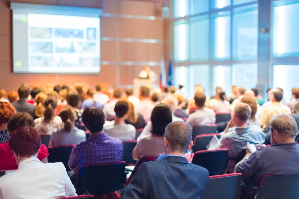 Auditório cheio com diversas pessoas sentadas assistindo a uma palestra ou apresentação corporativa. Ao fundo, uma tela de projeção exibe imagens e uma pessoa está em pé no púlpito conduzindo o evento.