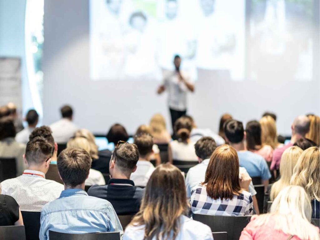Público participando de um seminário durante um evento corporativo. A imagem mostra diversos profissionais sentados, atentos a um palestrante que apresenta informações projetadas em uma tela.