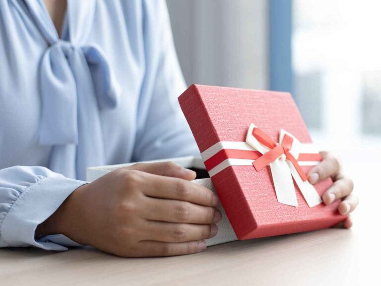 Cliente abrindo um brinde corporativo enviado pela empresa onde costuma adquirir produtos. Ela veste uma blusa azul com laço e segura uma embalagem vermelha.