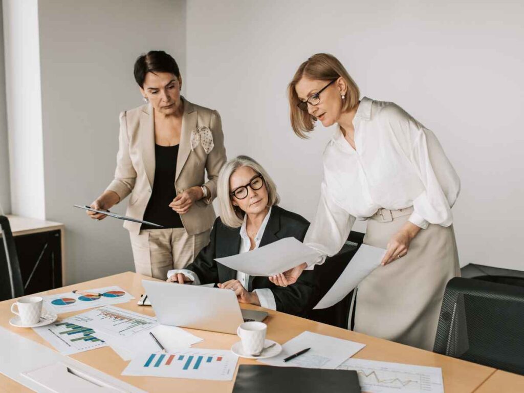 Dona de empresa sentada à mesa, cercada por papéis com gráficos, reunida com integrantes do RH para analisar o crescimento da empresa após a implementação de ações voltadas à melhoria do clima organizacional. As representantes do RH estão em pé ao seu lado, e uma delas apresenta um relatório impresso, destacando os resultados positivos da iniciativa.