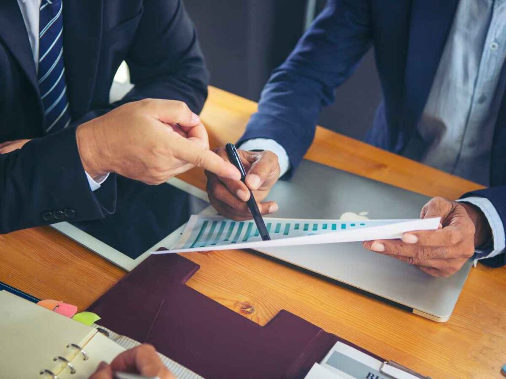 Mãos de dois sócios analisando o quanto eles podem gastar com brindes. Um deles, vestindo um terno azul escuro, aponta para uma folha com dados e gráficos que o outro, também de terno azul, segura. A cena representa a avaliação do orçamento da empresa para garantir que comprem brindes de qualidade.