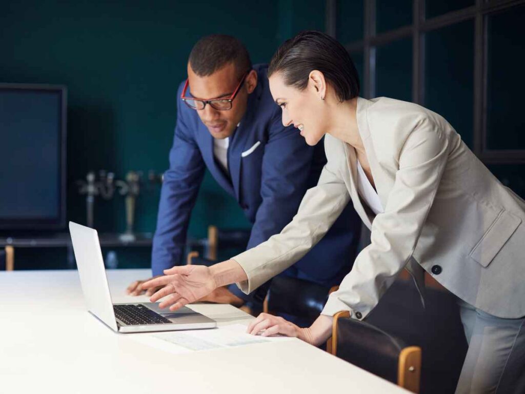 Colaboradora vestindo um conjunto creme apresenta para seu chefe, dono da empresa, algumas ideias e projetos. Eles estão em pé em frente a uma mesa branca, onde há um laptop branco. A colaboradora aponta para a tela do laptop enquanto explica, e seu chefe, vestindo um terno azul, observa atentamente e escuta com interesse.