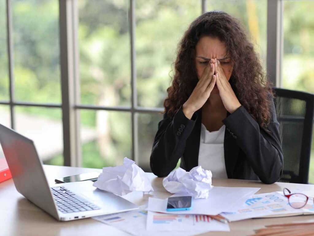 Colaboradora de uma empresa, com os cotovelos apoiados na mesa, segurando a cabeça com uma expressão de desânimo. Sobre a mesa, há inúmeros papéis amassados espalhados. Ela demonstra frustração por não se sentir valorizada, refletindo a falta de uma cultura de reconhecimento em sua empresa.