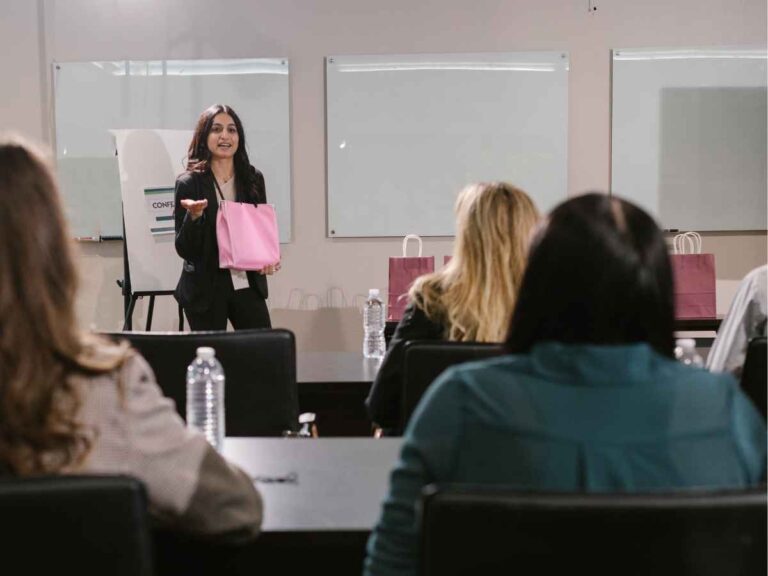 Mulher em pé segurando uma sacola de brinde que será entregue a uma das participantes de um evento corporativo. Ela está posicionada em frente a um grupo de executivas sentadas, que a escutam atentamente. Ao seu lado, há uma mesa com várias sacolas enfileiradas.