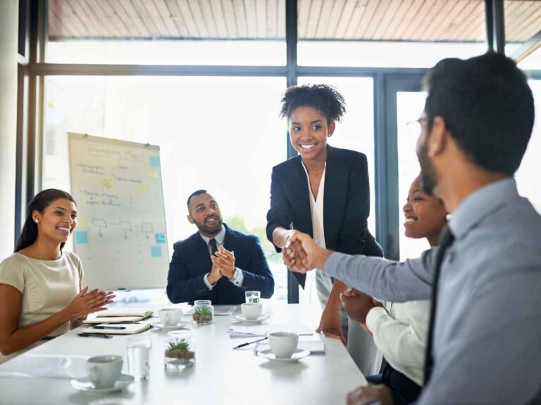 Donos de uma empresa com uma cultura de reconhecimento eficiente, reunidos em torno de uma mesa para parabenizar uma colaboradora por seu empenho e resultados alcançados. Um dos donos aperta a mão da colaboradora em sinal de reconhecimento e valorização.
