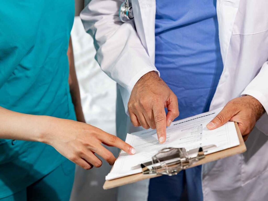 Médico e profissional da enfermagem analisando juntos uma prancheta com documentos. Ambos apontam para o mesmo ponto na folha, em um momento de troca de informações e colaboração.