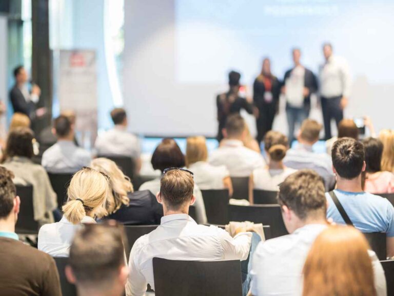 Público assistindo atentamente a uma apresentação durante um evento corporativo. A imagem mostra uma plateia composta por profissionais sentados, enquanto palestrantes se apresentam no palco ao fundo.