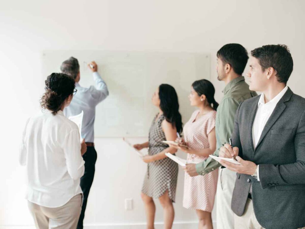 Dono de empresa escrevendo em uma lousa as estratégias de reconhecimento que serão implementadas, refletindo a adoção de uma cultura de valorização na empresa. Os colaboradores observam atentamente e fazem anotações, demonstrando engajamento com as novas iniciativas.