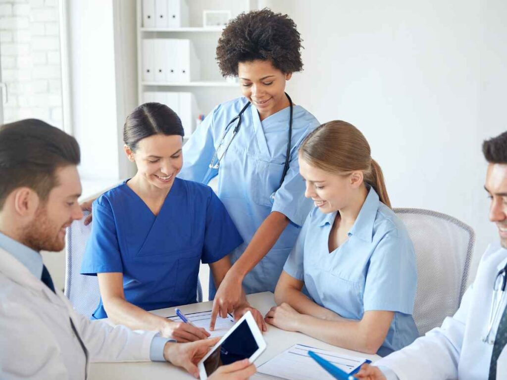 Equipe multiprofissional da área da saúde reunida ao redor de uma mesa, sorrindo e analisando documentos e um tablet. A imagem retrata um momento onde os integrantes do SESMT analisam o impacto que suas ações tiveram na empresa.