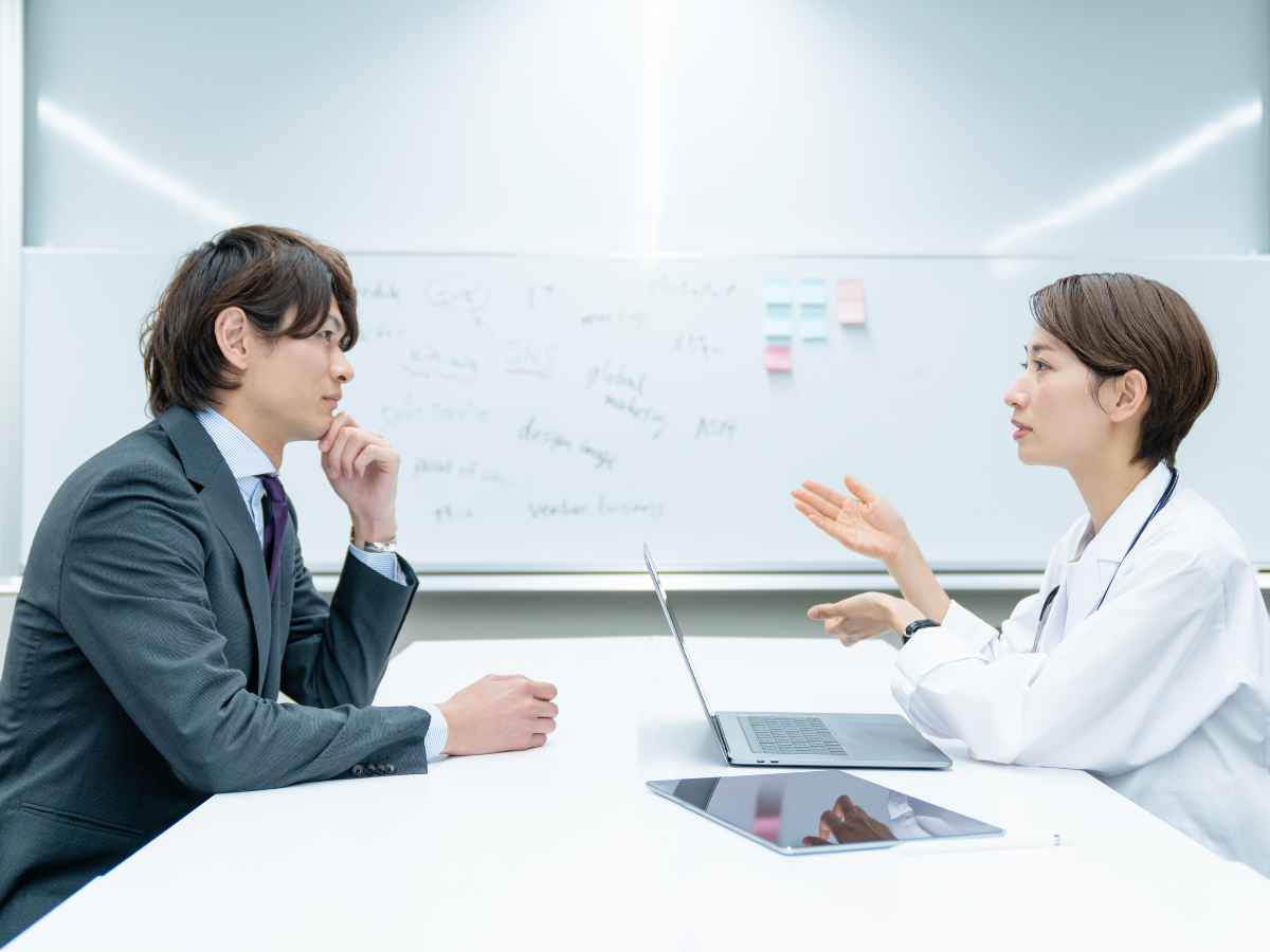 Médica do trabalho conversando com um colaborador em uma sala corporativa. Sentados frente a frente, ela explica algo com gestos enquanto utiliza um notebook, e ele ouve com atenção, com expressão pensativa. Ao fundo, há um quadro branco com anotações.