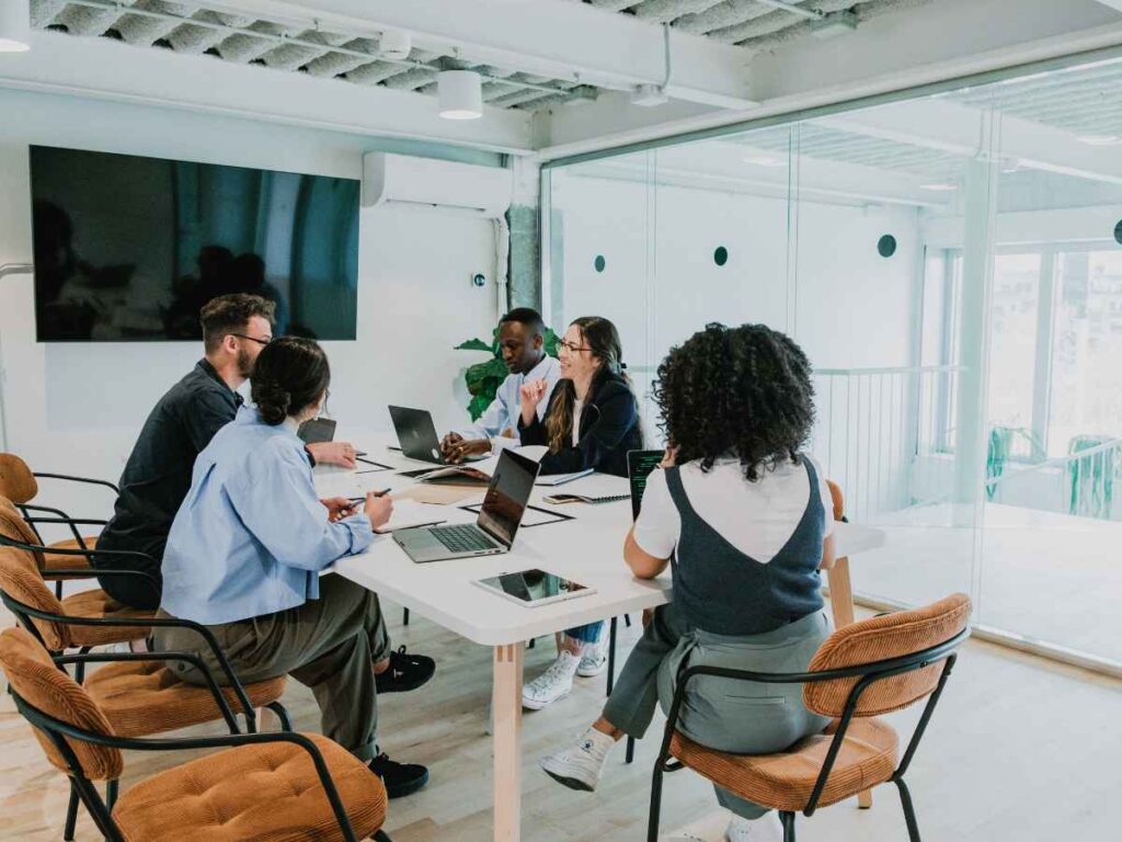 Equipe de 5 pessoas reunidas em torno de uma mesa, engajada no planejamento de um evento corporativo. Com laptops, anotações e materiais de apoio espalhados, os integrantes discutem ideias, definem estratégias e colaboram entre si.