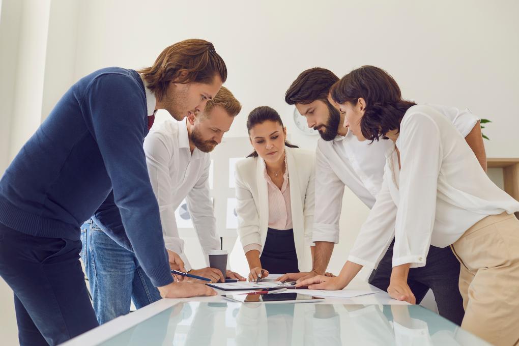 equipe empresarial de 5 pessoas em uma reunião, 2 mulheres e 3 homens em pé em volta de uma mesa de vidro
