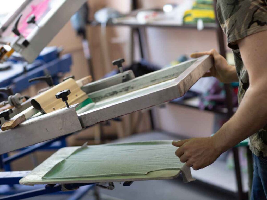 Profissional operando máquina de silk screen em processo de personalização de camiseta verde, demonstrando a aplicação manual de tinta sobre tecido por meio de tela serigráfica.