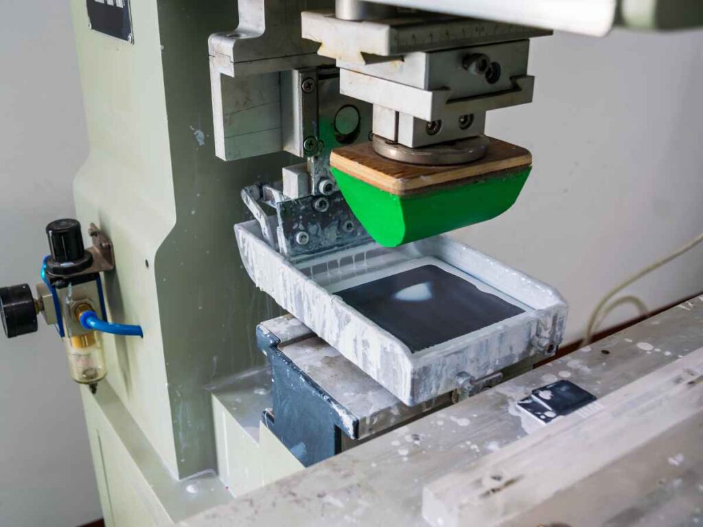 Máquina de tampografia em funcionamento, com almofada de silicone coletando tinta de um clichê para transferir a imagem para objetos, representando o processo de personalização de brindes.