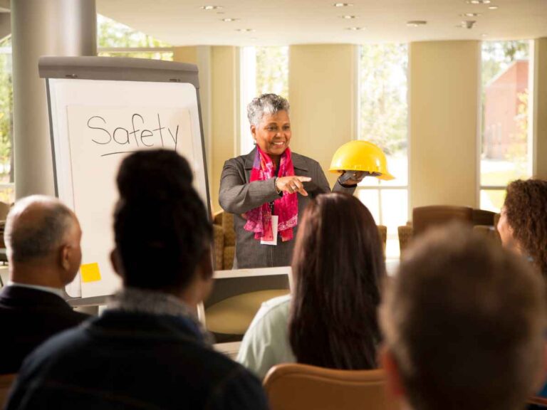 Palestrante segurando um capacete amarelo enquanto conversa com um grupo de pessoas sentadas, em frente a um quadro com a palavra “Safety”; a imagem representa uma equipe engajada em uma campanha de segurança no trabalho promovida pelo SESMT.