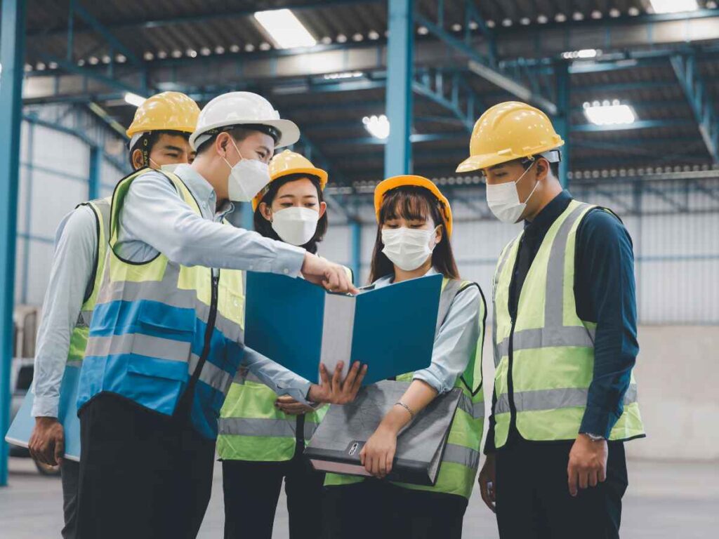 Grupo de profissionais com coletes refletivos, capacetes e máscaras faciais analisa um documento em ambiente industrial; cena representa uma reunião conduzida pelo SESMT.