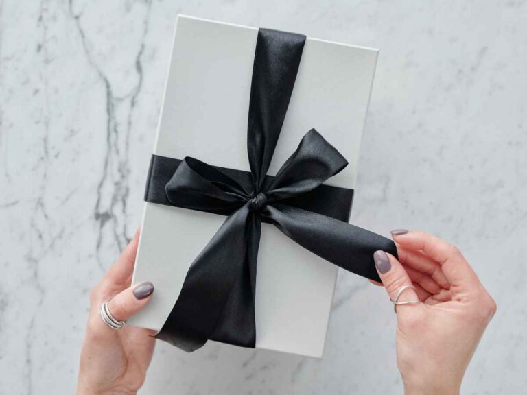 Mãos femininas abrindo uma caixa de presente branca com laço preto de cetim, em fundo de mármore claro.