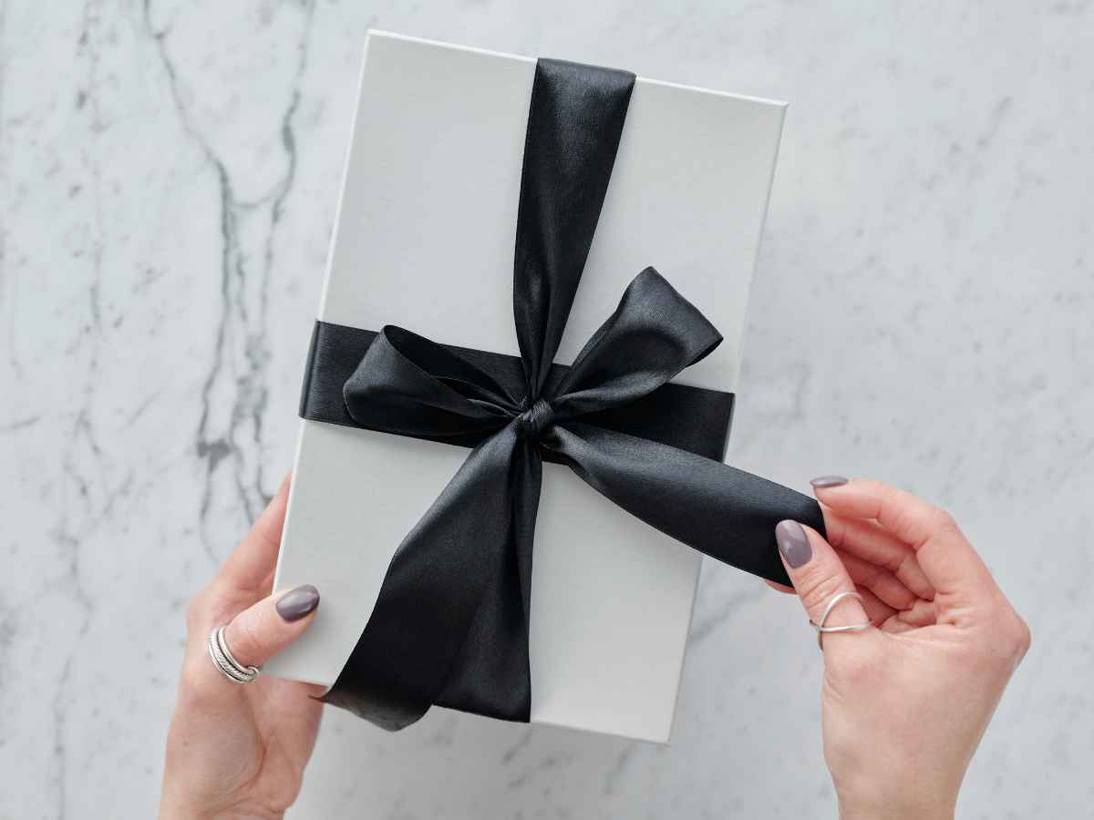 Mãos femininas abrindo uma caixa de presente branca com laço preto de cetim, em fundo de mármore claro.