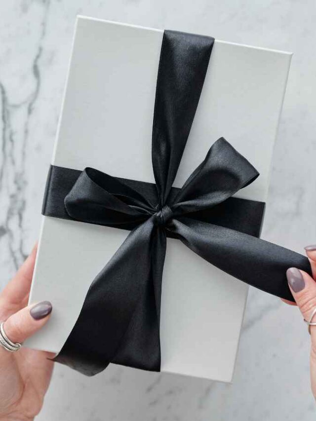 Mãos femininas abrindo uma caixa de presente branca com laço preto de cetim, em fundo de mármore claro.