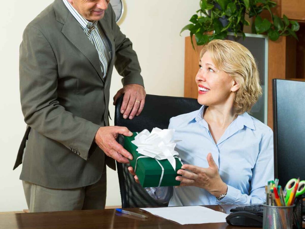 Homem entrega presente embrulhado com laço branco para secretária no escritório, simbolizando reconhecimento e valorização profissional.