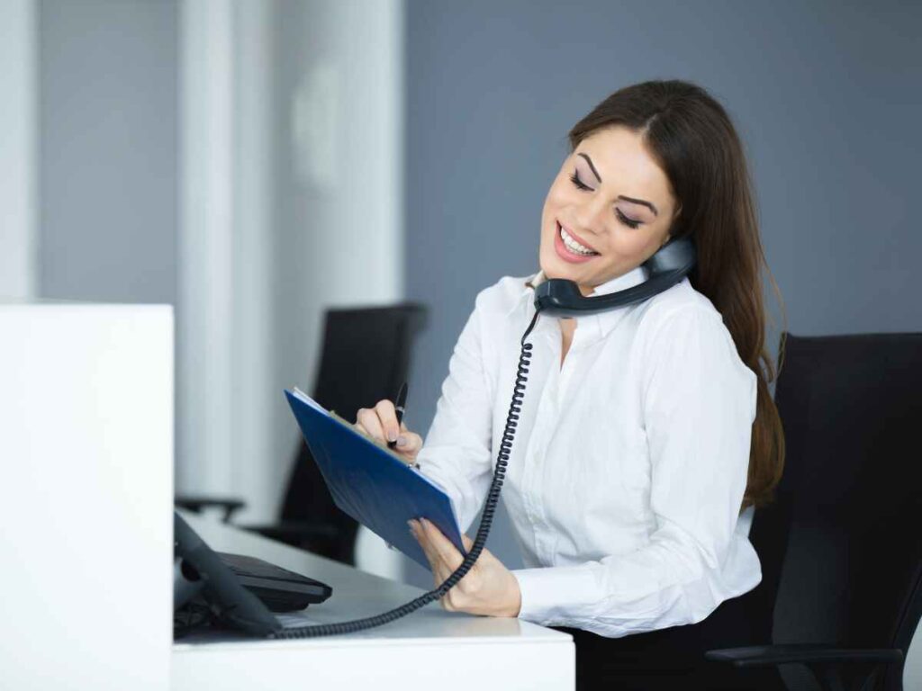 Secretária sorridente atendendo ligação telefônica enquanto anota informações em uma prancheta azul, sentada em sua mesa de trabalho no escritório.
