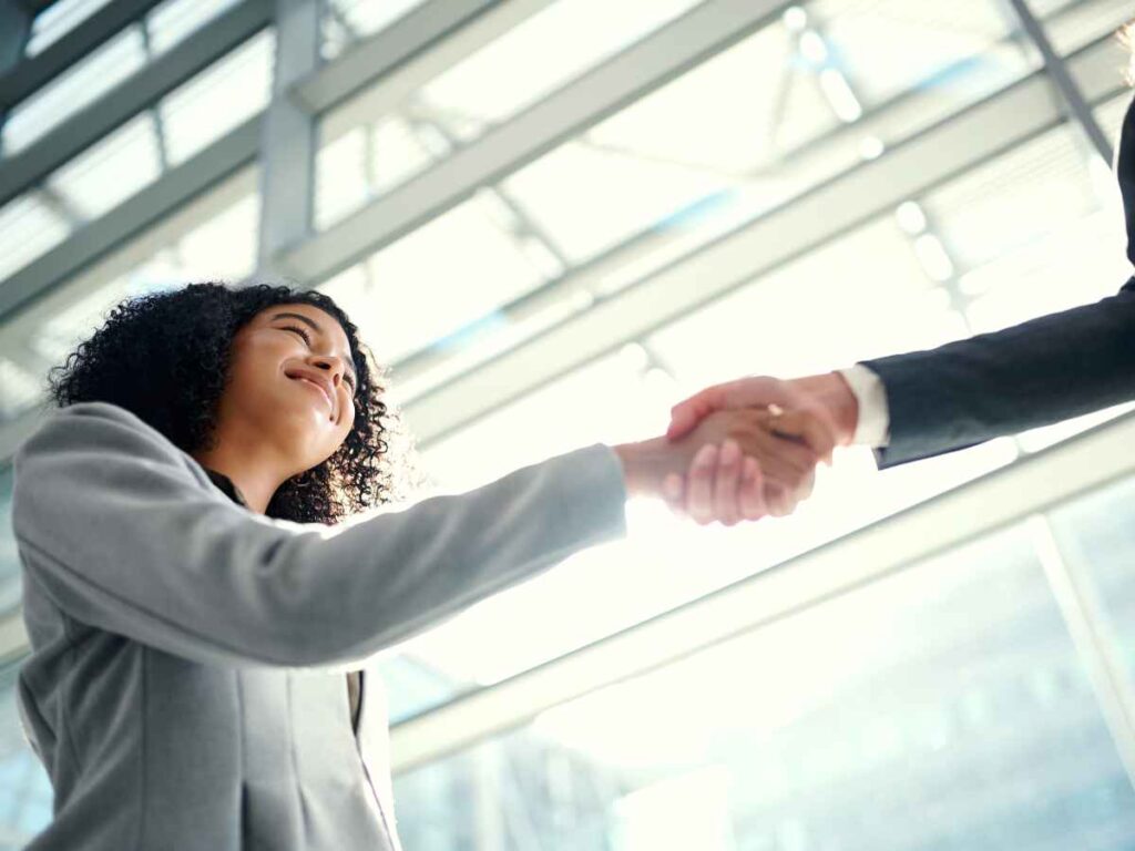 Imagem de duas pessoas em trajes formais apertando as mãos em um ambiente corporativo moderno, com vidros e estrutura metálica ao fundo.