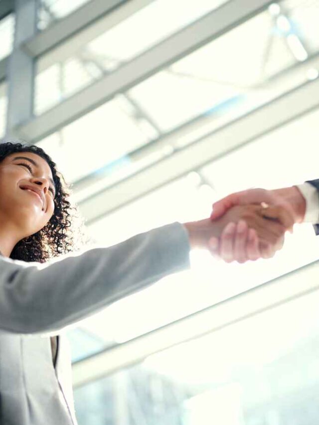 Imagem de duas pessoas em trajes formais apertando as mãos em um ambiente corporativo moderno, com vidros e estrutura metálica ao fundo.