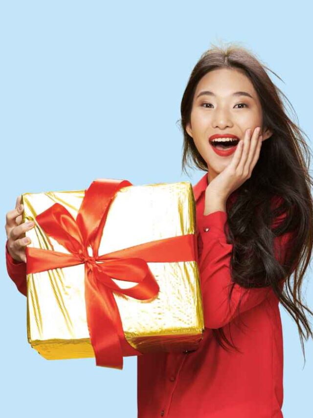 Mulher jovem sorridente, vestindo blusa vermelha, segura um presente dourado com laço vermelho e expressa surpresa e alegria. Fundo azul claro.