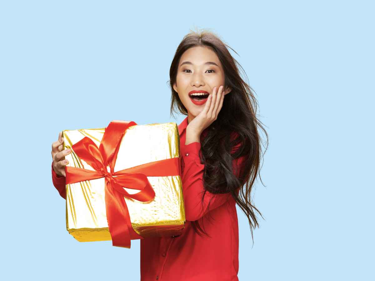 Mulher jovem sorridente, vestindo blusa vermelha, segura um presente dourado com laço vermelho e expressa surpresa e alegria. Fundo azul claro.